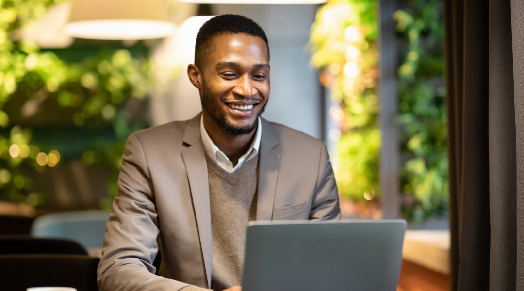 Man in suit jacket sitting behind a laptop, facing screen, and smiling.