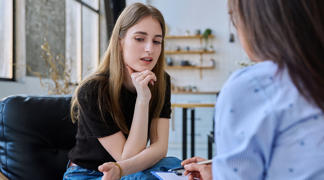 One woman, with long hair and hand on chin, facing another person. Engaged in conversation.