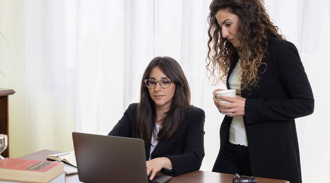 Two women. One is standing and holding a coffee mug. The other is sitting. They are both looking at a shared computer screen.